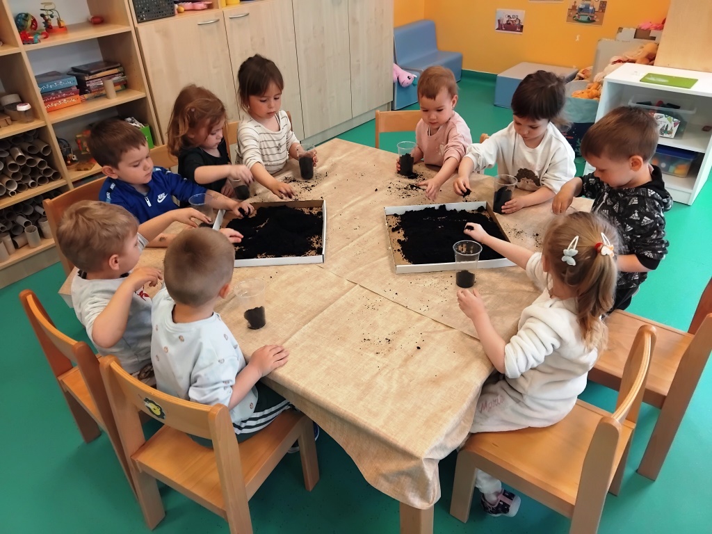 Djeca u vrtiću sjede oko stola i istražuju zemlju, pune male čaše i posude dok sudjeluju u zajedničkoj aktivnosti.