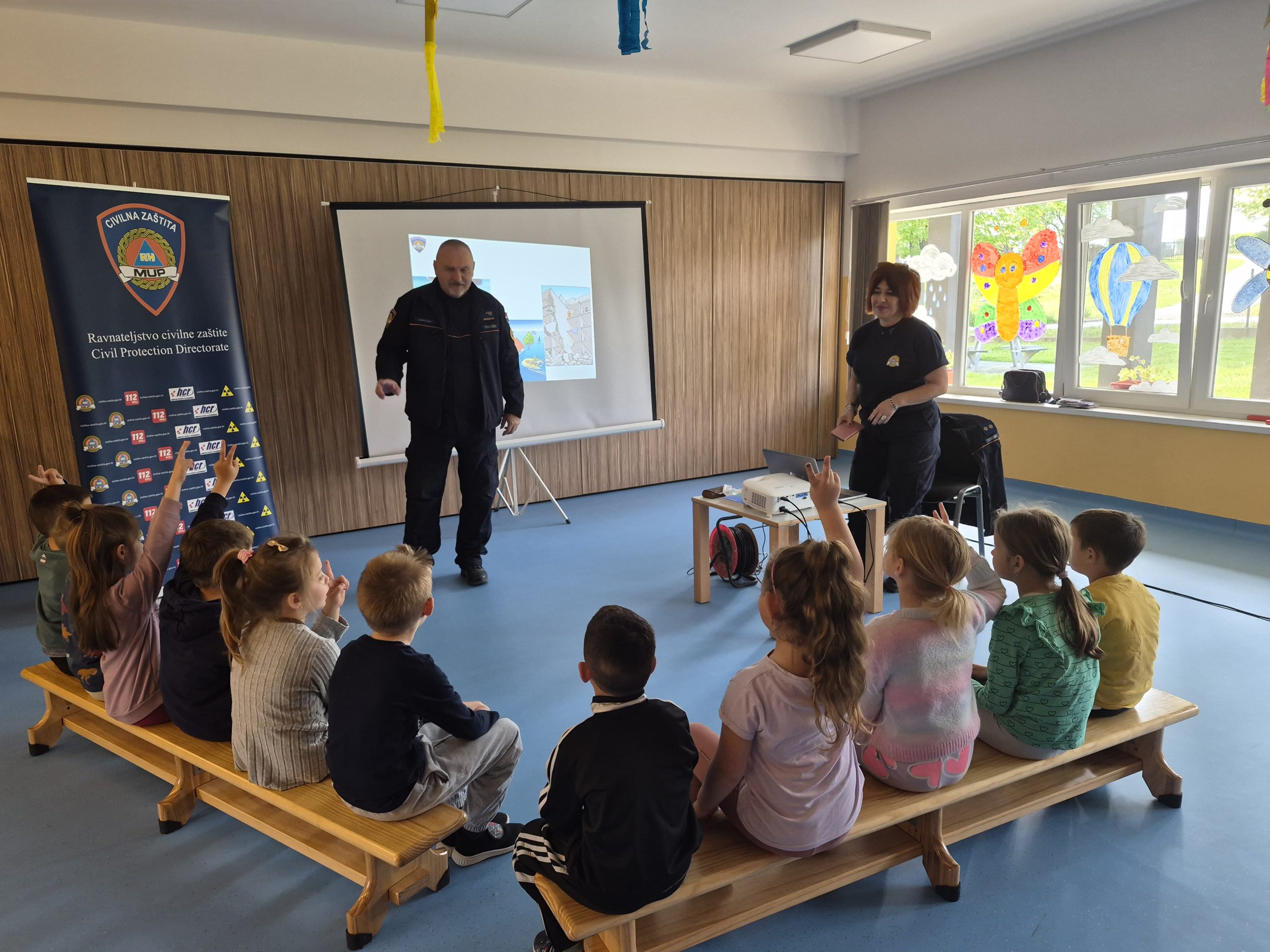Djeca sjede na klupama u dvorani i podižu ruke dok ih dvoje djelatnika civilne zaštite podučava uz projektor i prezentaciju.