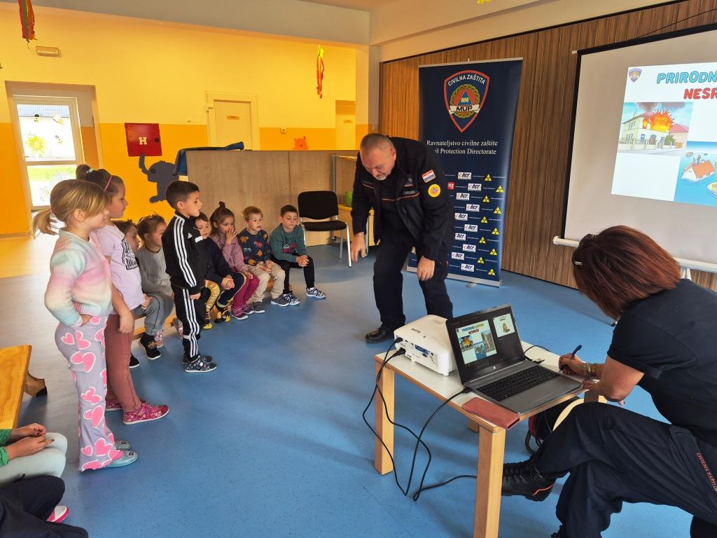 Djeca stoje u polukrugu i prate demonstraciju djelatnika civilne zaštite, dok djelatnica upravlja prezentacijom na laptopu i projektoru.