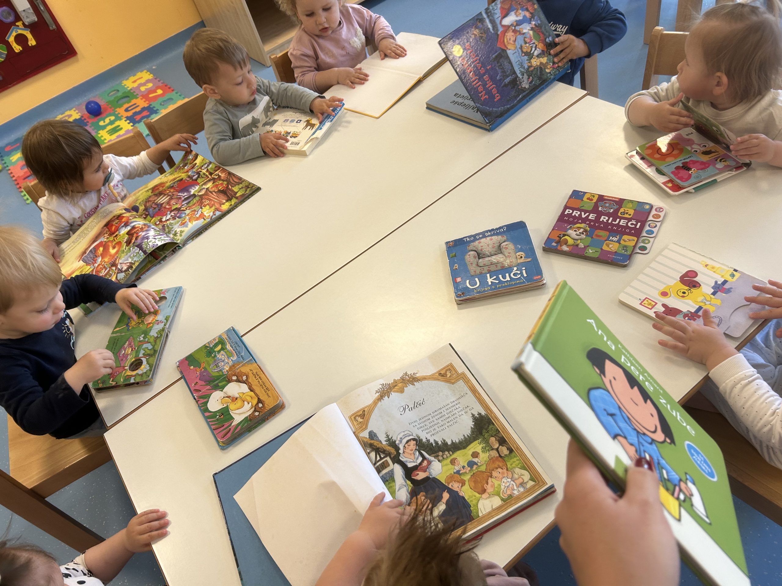 Grupa djece sjedi za stolom, okružena knjigama, razgovaraju i dijele priče.