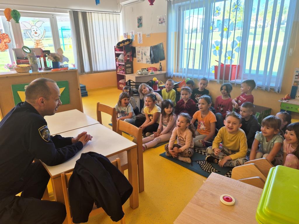 Policijski službenik sjedi za stolom. ispred njega na podu sjede djeca i slušaju edukaciju. Atmosfera je vesela. fotografija je slikana iz suprotnog kuta.