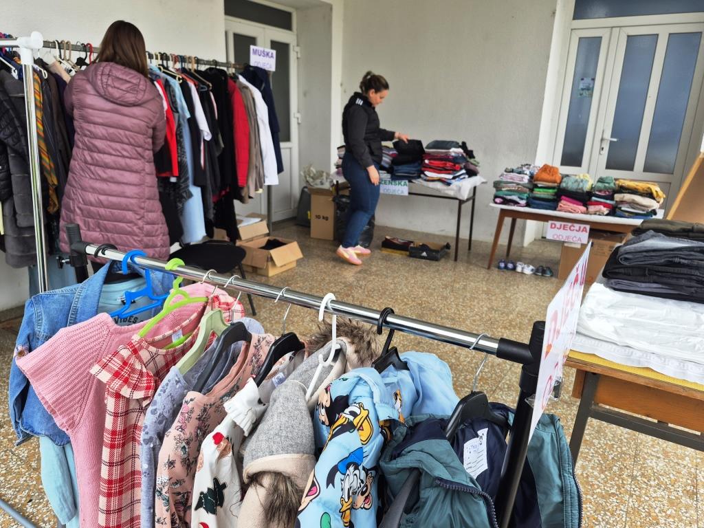 Štand s doniranom odjećom – dječja garderoba na vješalicama u prvom planu, dok dvije žene razvrstavaju odjeću na stolovima u pozadini