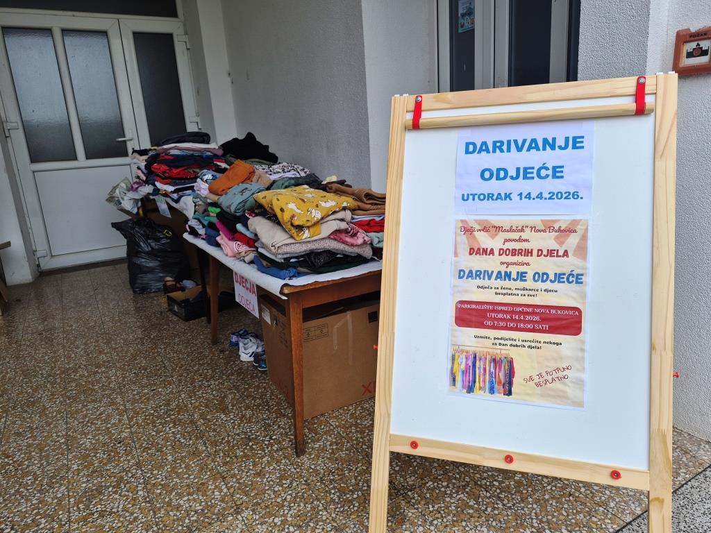 Stolovi prekriveni složenom odjećom za donaciju uz plakat ‘Darivanje odjeće’ s datumom događaja, postavljen na stalku ispred ulaza u zgradu.