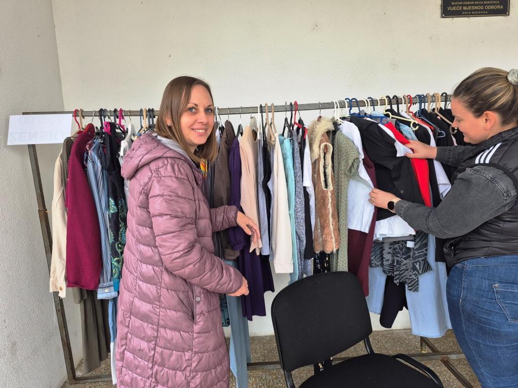 Odjeća na vješalici, a u prvom planu ispred odjeća se nalaze dvije ženske osobe koje pregledavaju odjeću.