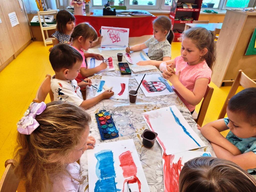 Skupina djece u vrtićkoj sobi sjedi i stoji oko stola te slika vodenim bojama. Na papirima su naslikane crvena i plava polja  inspirirana bojama hrvatske zastave. Djeca surađuju, dijele pribor za slikanje i koncentrirano rade na likovnom zadatku.