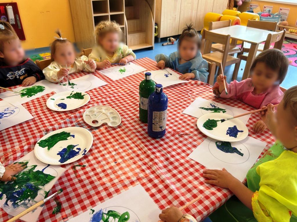 Dan-planeta-Zemlje-II - Skupina male djece sjedi oko stola i slika zelenom i plavom bojom na papirima, koristeći kistove i tanjuriće s bojom.