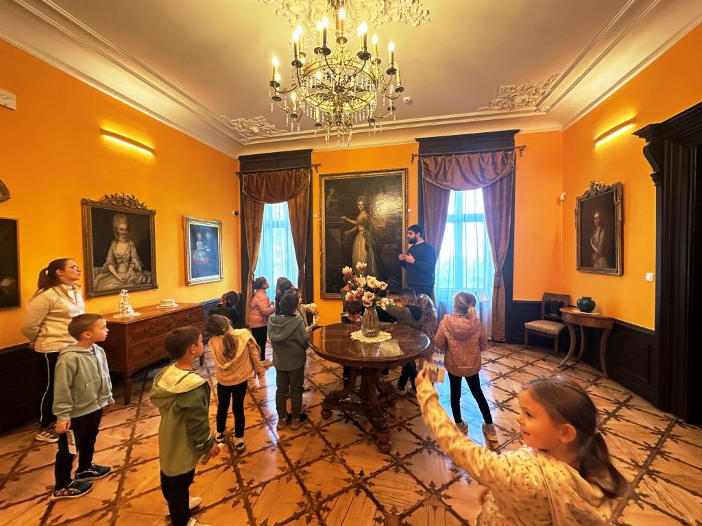 Izlet-skupine-Tockice-I - Djeca se nalaze u unutrašnjosti dvorca gdje stoje i slušaju predavanje kustosa. U prostoru se nalaze slike obitelji Pejačević, stari namještaj te velika dva prozora sa zavjesama.