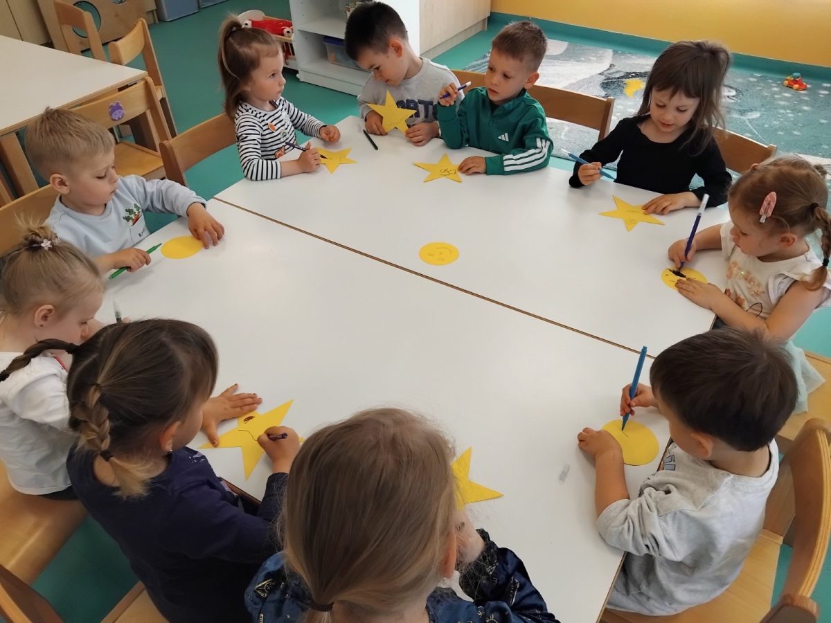 Noc-knjige-I - Djeca sjede oko bijelog stola i crtaju markerima po žutim izrezanim zvijezdama i mjesecima. Učionica je svijetla, stvara veselu, kreativnu atmosferu.