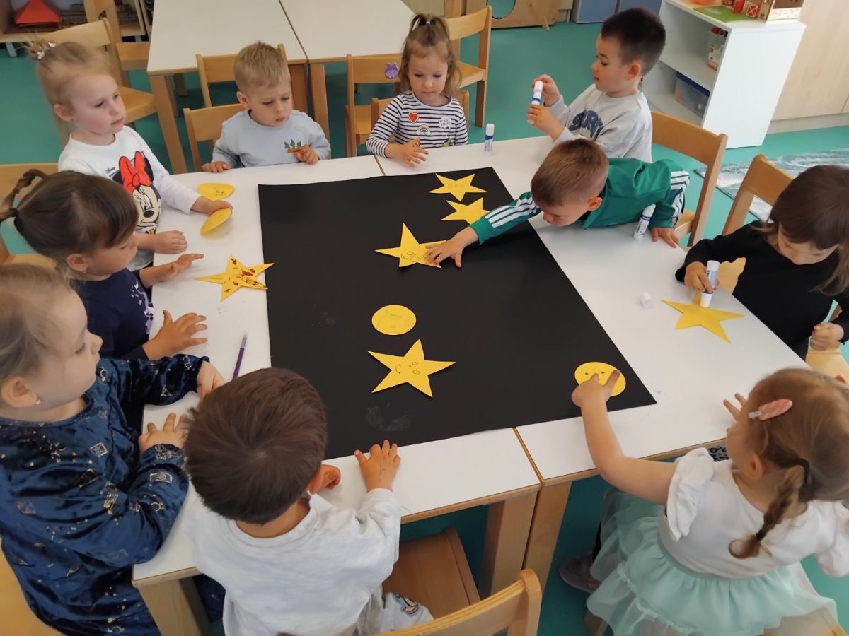 Noc-knjige-II - Djeca sjede oko bijelog stola i rade na kreativnom projektu. Ljepilom lijepe žute oblike zvijezda i mjeseca na veliki crni plakat. Djeca su angažirana i surađuju, nasmijana su i koncentrirana na svoj umjetnički rad. Radnja se odvija u prostoriji s drvenim stolicama i zelenim podom.