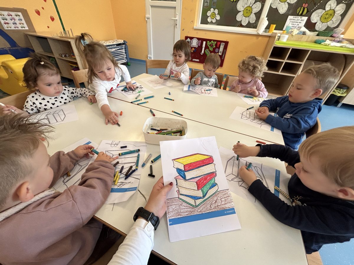 Noc-knjige-IV - Grupa male djece sjedi oko stola u učionici i boji slike knjiga bojicama. Čine se usredotočenima i angažiranima u svojoj aktivnosti.