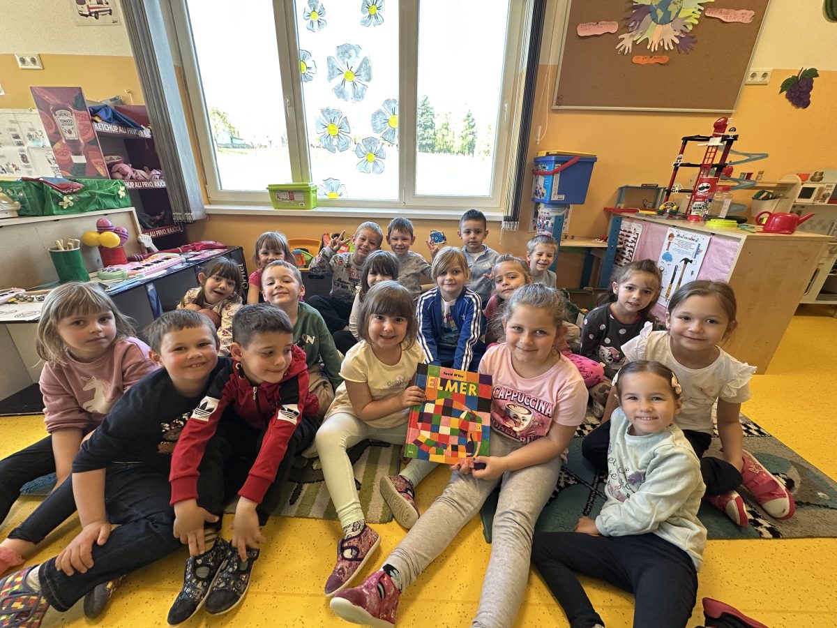 Noc-knjige-V - Grupa nasmijane djece sjedi na podu prostorije držeći šarenu knjigu pod nazivom Elmer. Prostorija je svijetla i uređena edukativnim stilom.