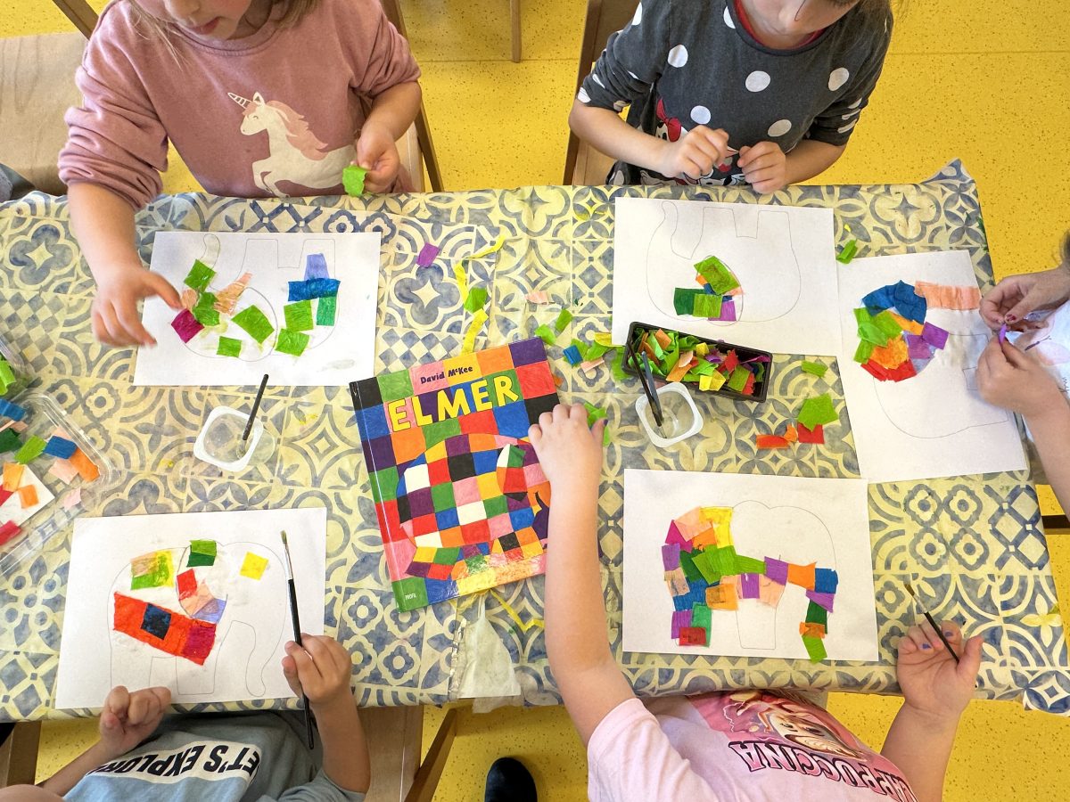 Noc-knjige-VI - Djeca za stolom izrađuju šarene papirnate kolaže slona Elmera. Vidljivi su knjiga o Elmeru i kvadratići od obojenog papira.