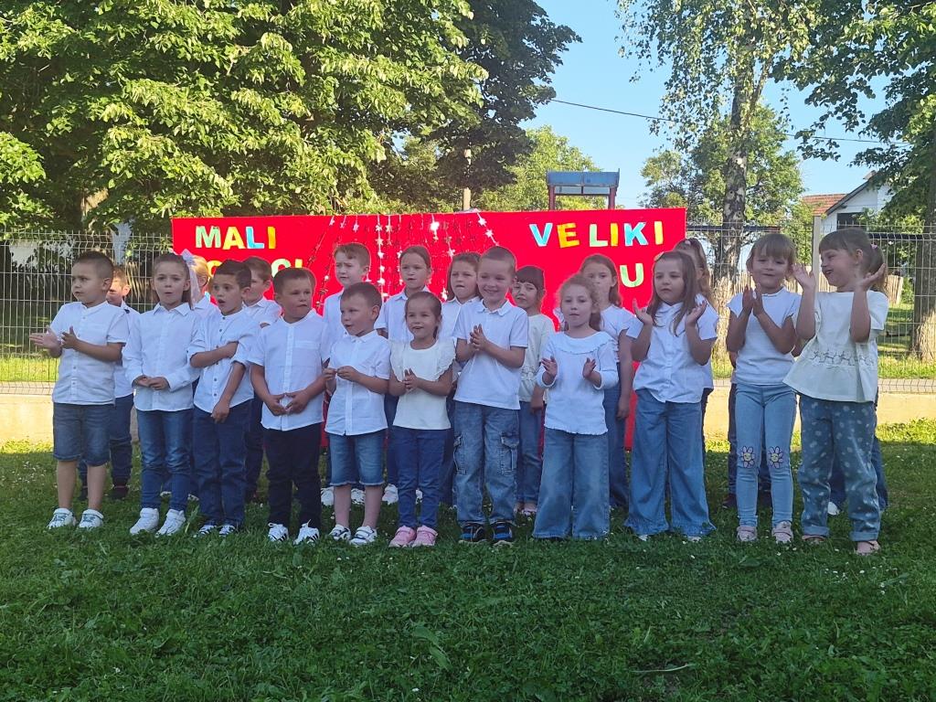 Grupa djece stoji ispred znaka, smiješeći se i pozirajući za fotografiju. 