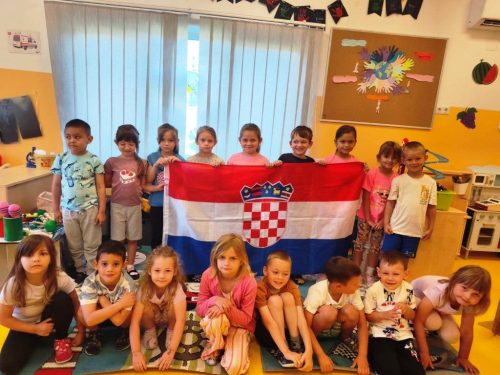 Grupa djece u sobi pozira držeći hrvatsku zastavu, s igračkama i šarenim ukrasima u pozadini.