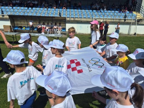 Grupa djece u bijelim kapama i majicama drži veliku zastavu s olimpijskim krugovima i natpisom HRVATSKA na sportskom terenu, dok u pozadini sjede gledatelji.