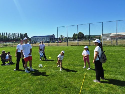 Djeca i odrasli u bijelim majicama i kapama sudjeluju u aktivnosti na otvorenom na travnatoj površini pod vedrim plavim nebom. Na tlu su vidljivi sportska oprema i žuta linija.