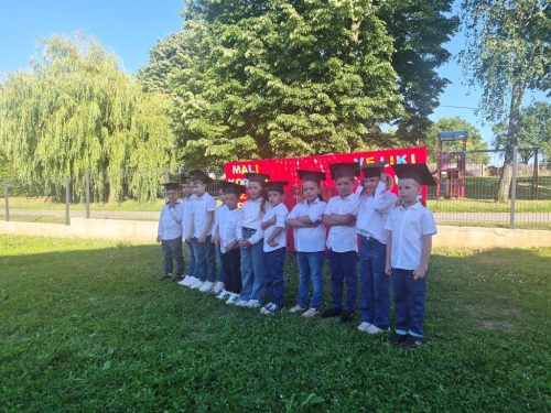 Grupa djece u bijelim majicama i maturantskim kapama stoji u redu na travi ispred drveća i crvenog znaka vani.